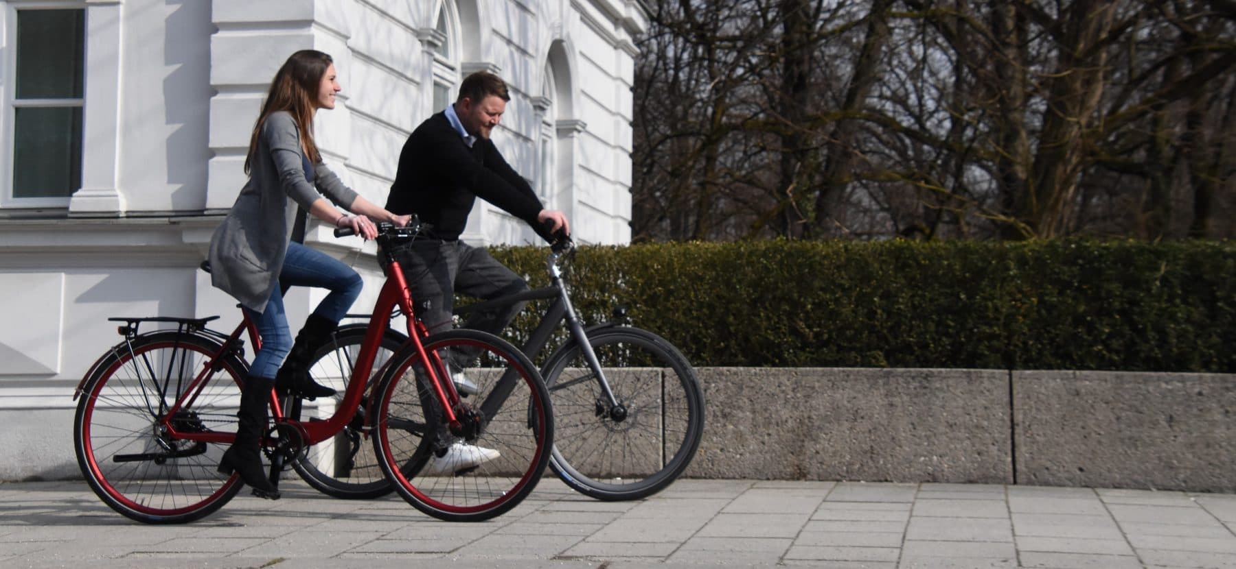 Zwei Menschen fahren auf Leihrädern an einem weißen Altbau in München vorbei